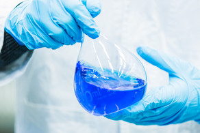 Zwei Hände in blauen Gummihandschuhen halten einen Messkolben, der mit blauer Flüssigkeit gefüllt ist.